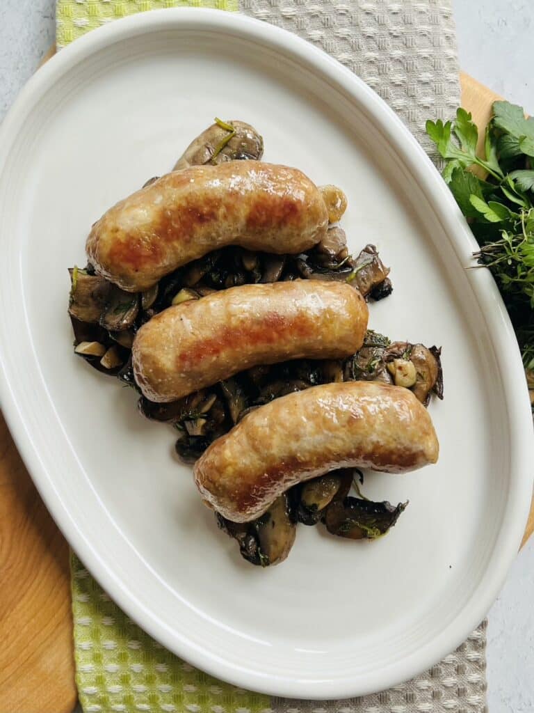 Oven baked Italian sausage over sautéed mushrooms on white platter.