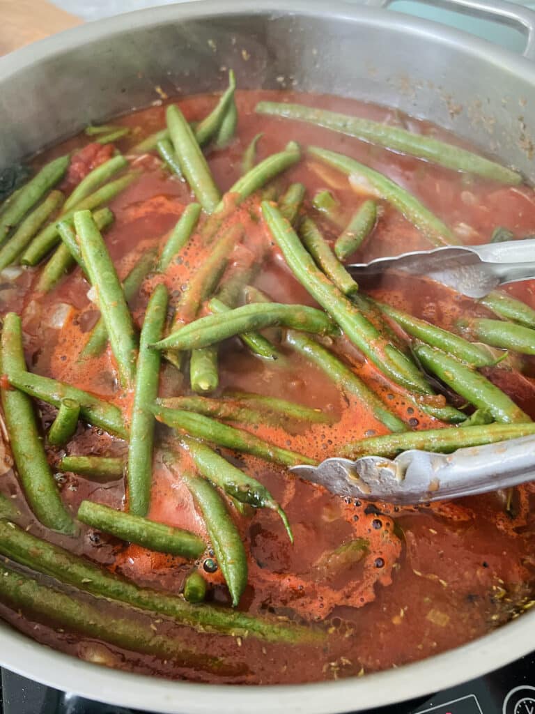 Green beans in tomato mixture in pan with tongs.