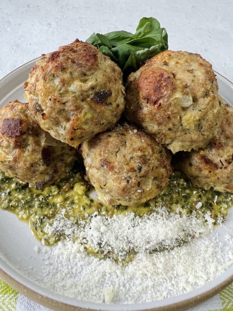 Styled close up of cooked turkey meatballs with pesto on top of pesto on plate, garnished with grated cheese and whole basil leaves.