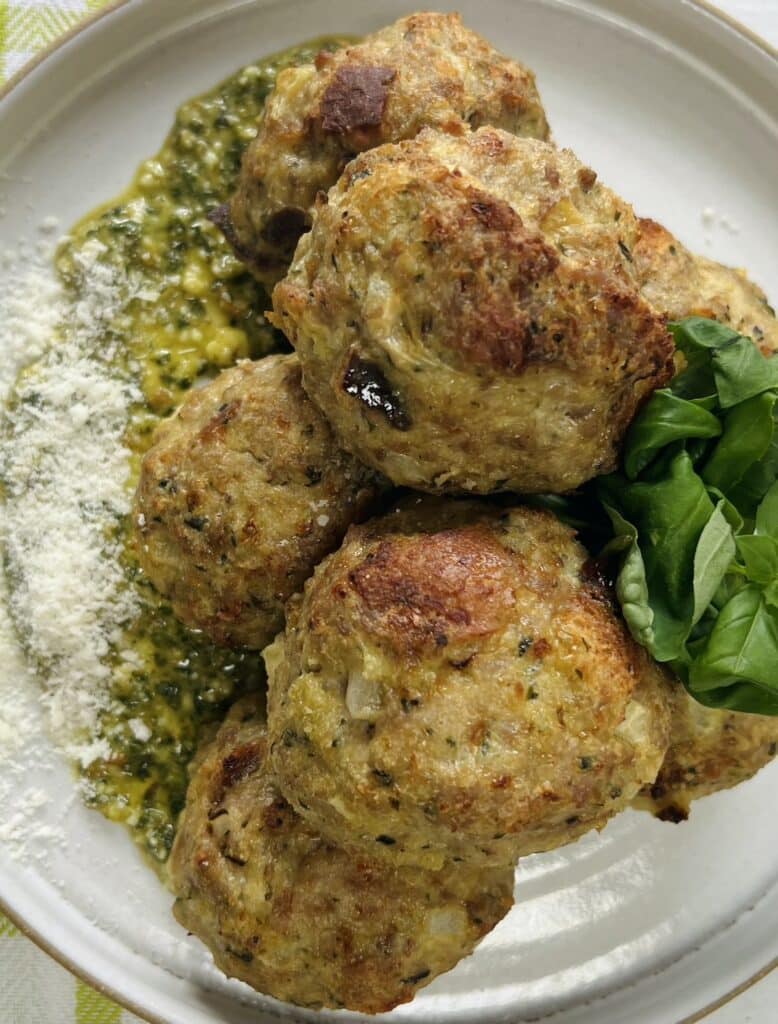 Overhead view of styled close up of cooked turkey meatballs with pesto on top of pesto on plate, garnished with grated cheese and whole basil leaves.