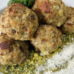 Styled close up of cooked turkey meatballs with pesto on top of pesto on plate, garnished with grated cheese and whole basil leaves.