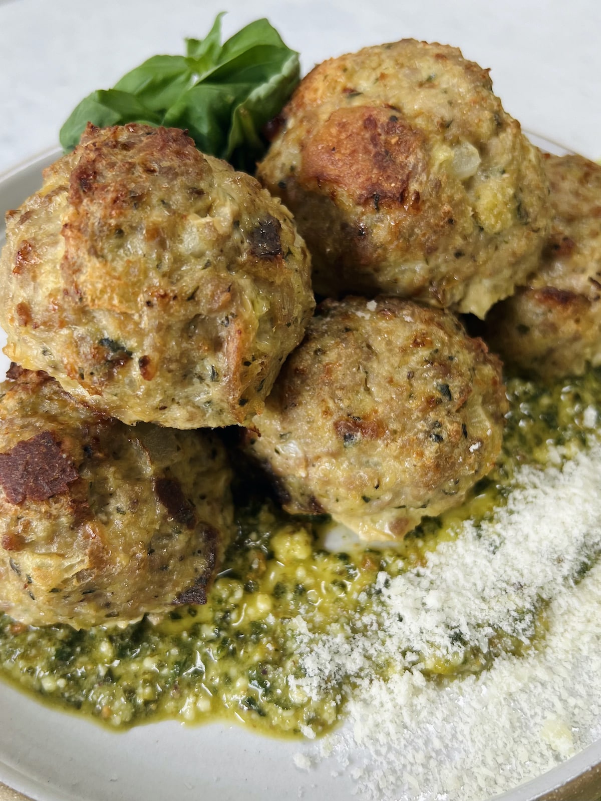 Styled close up of cooked turkey meatballs with pesto on top of pesto on plate, garnished with grated cheese and whole basil leaves.