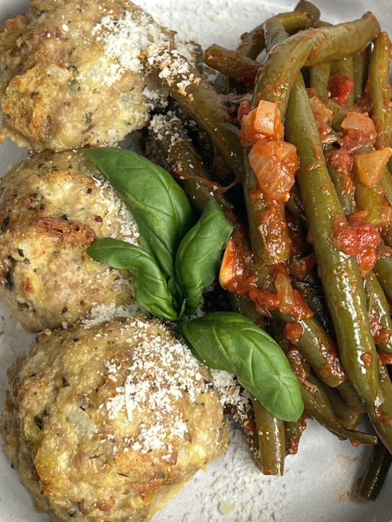 Overhead view of three cooked turkey meatballs with pesto on plate with green beans in tomato sauce and basil garnish.