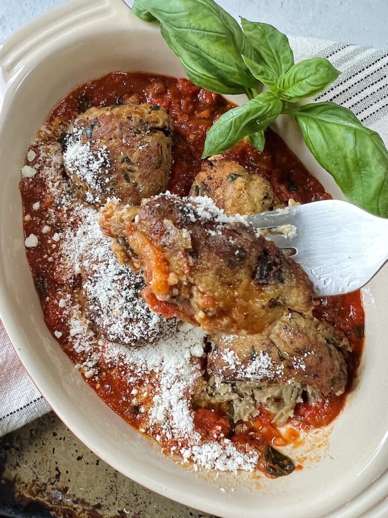 Close-up of forkful of polpette di melanzane over oval dish with basil garnish and grated cheese.