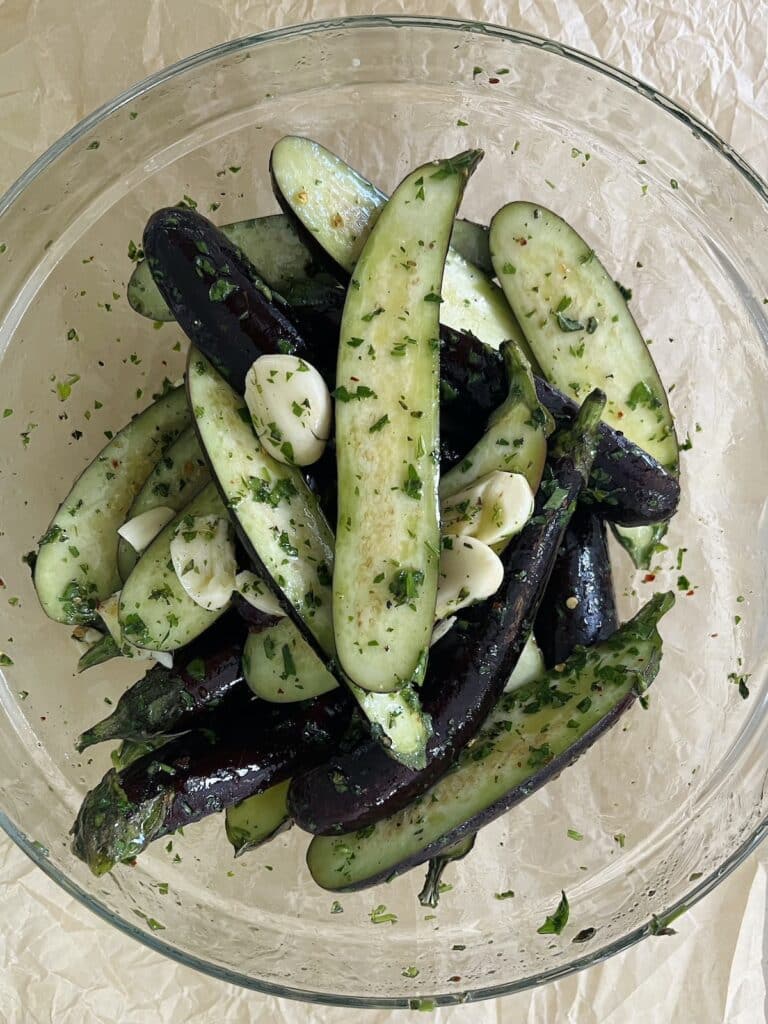 Bowl of halved eggplants with smashed garlic cloves and herb olive oil mixture.
