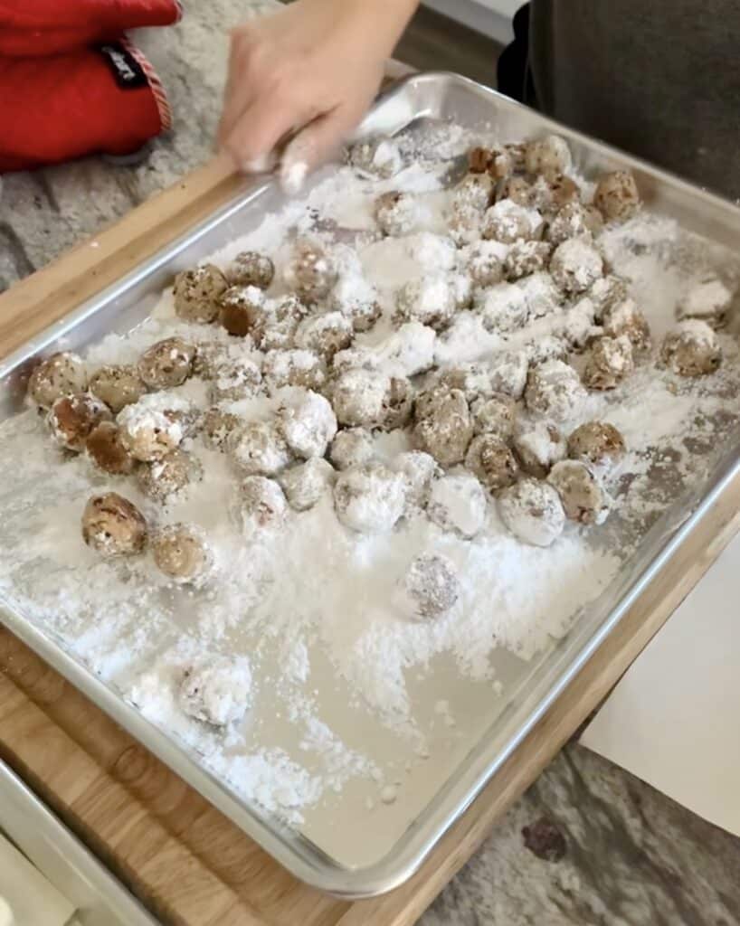 Buttery Pecan Snowball Cookies being tossed in powdered sugar on sheet pan.