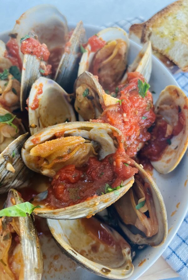 Clams posillipo in red sauce in white bowl topped with chopped parsley and toasted bread in background.