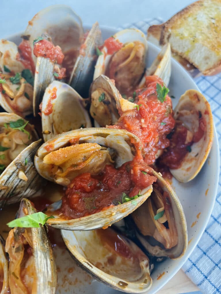 Clams posillipo in red sauce in white bowl topped with chopped parsley and toasted bread in background.