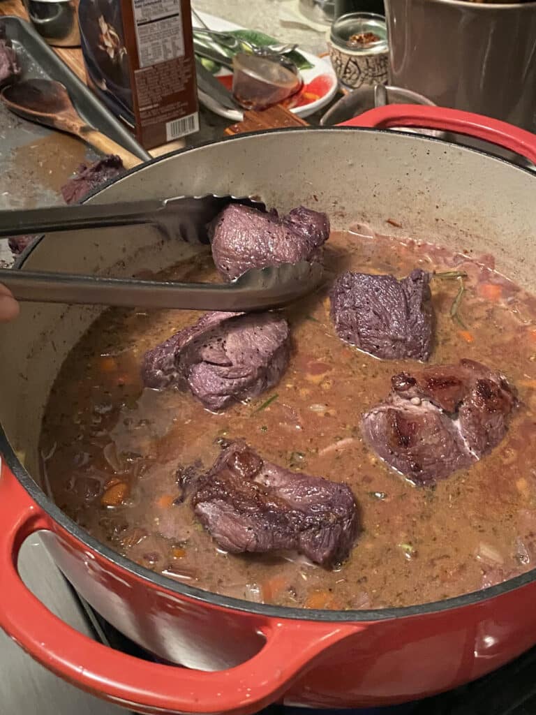 Adding seared pieces of boar back into the Dutch oven, into the braising mixture.