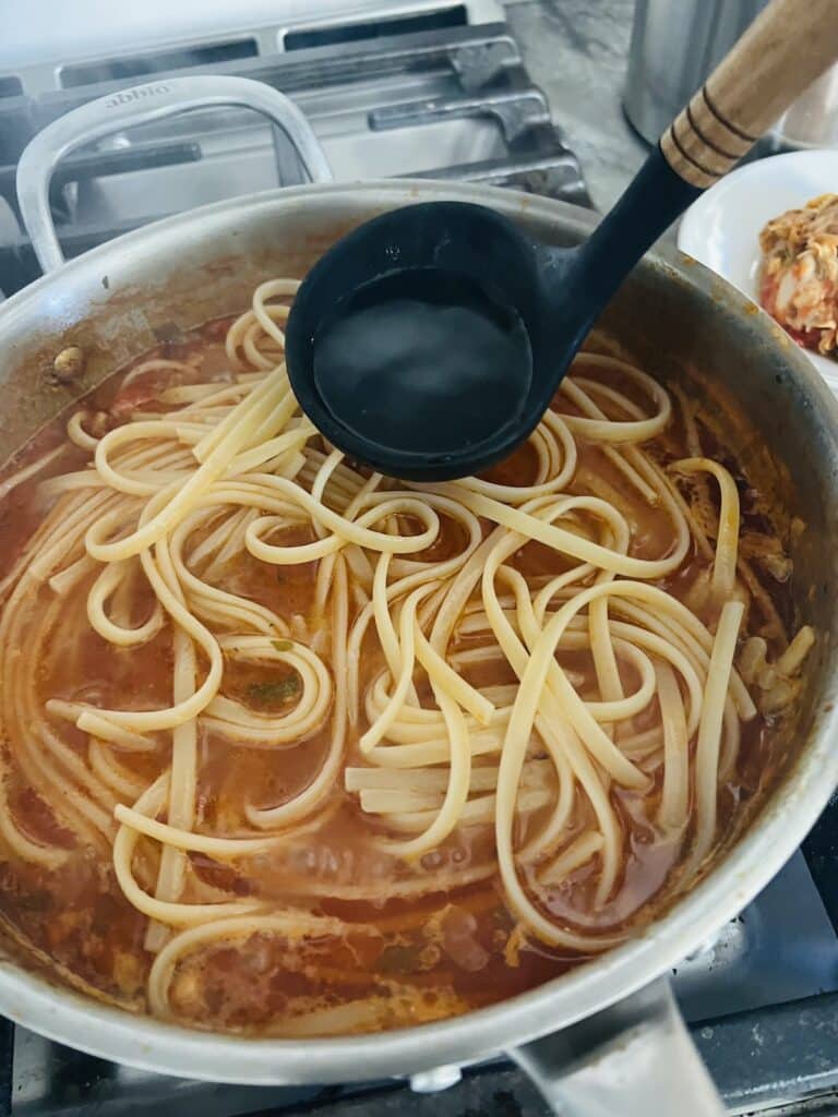 Adding a ladle of pasta water to pasta in sauce.