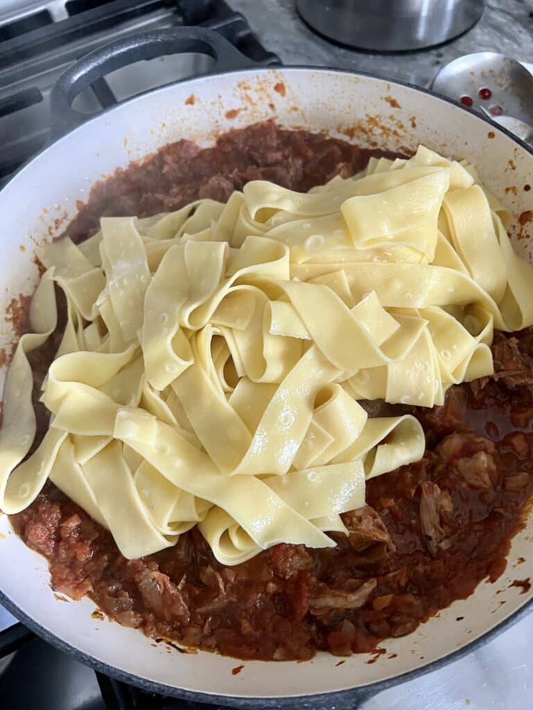 Cooked pasta added to pork ragu recipe in pan.