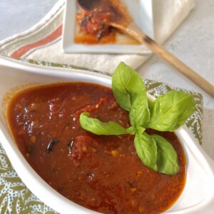 Finished sauce from frozen tomatoes in gravy boat with basil garnish with wooden spoon in background.