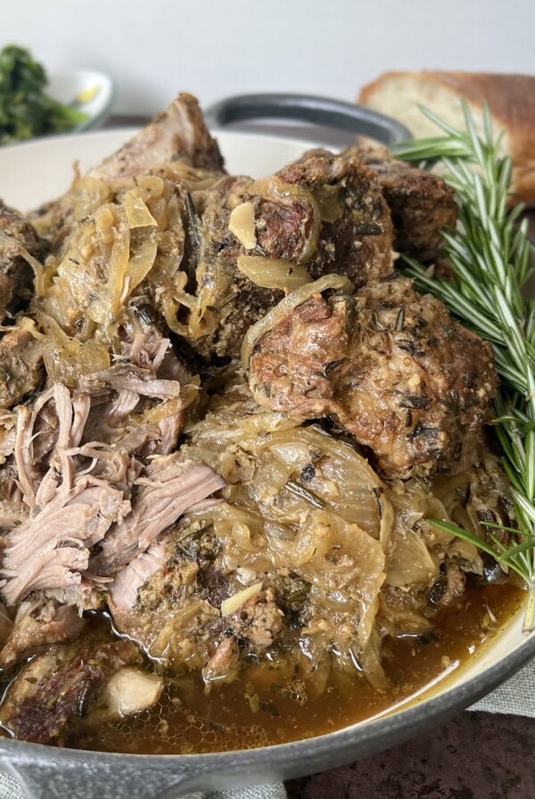 Side view of Italian Pulled Pork with onions and rosemary sprigs styled in gray pan with a platter of sautéed broccoli rabe and bread in the background.