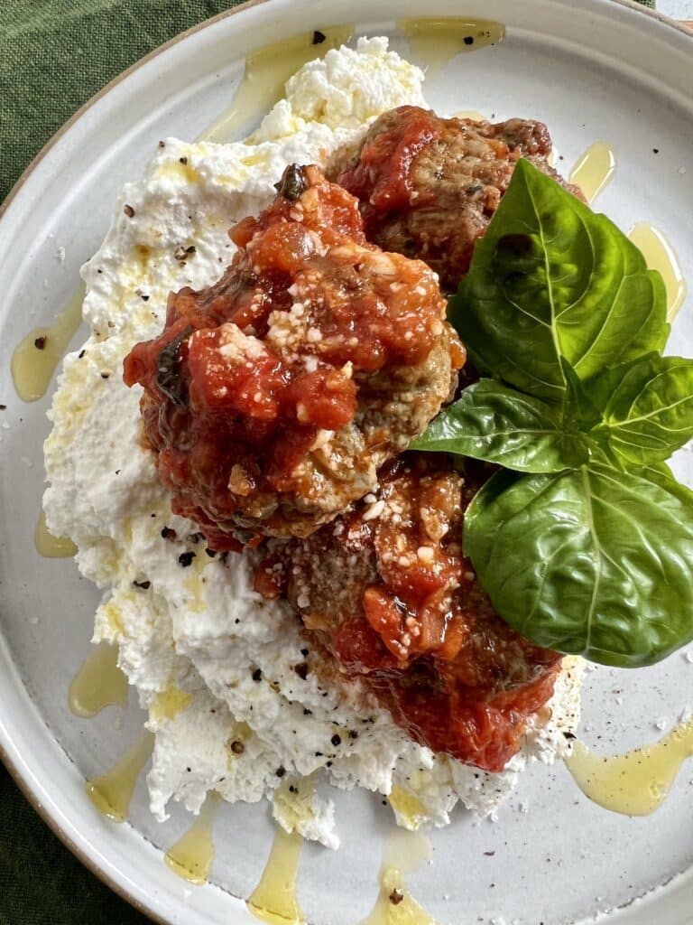 Pan fried meatballs in tomato sauce with basil garnish over a bed of creamy ricotta cheese.