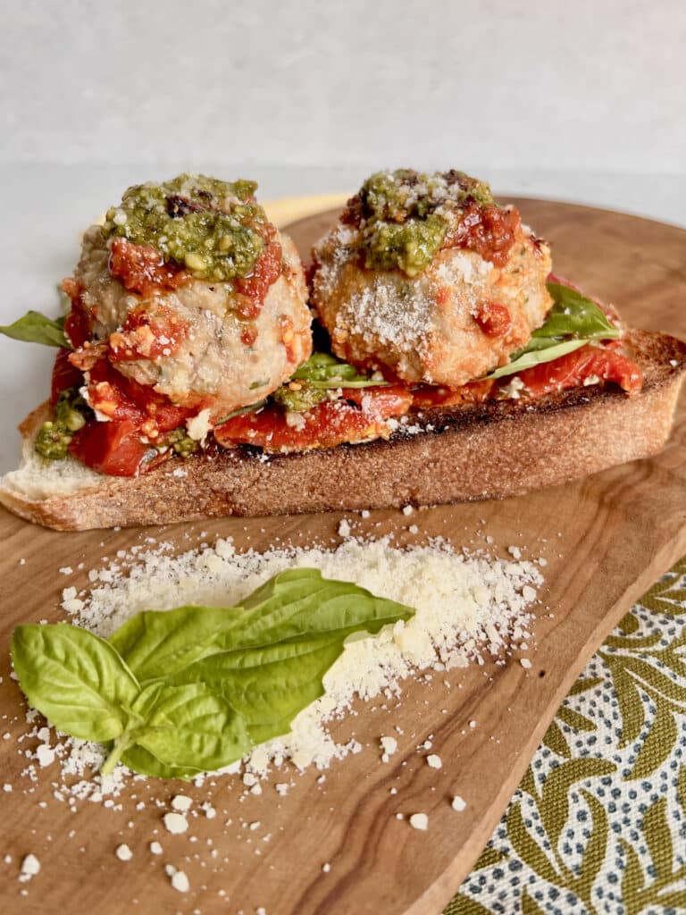 Meatballs No Bread Crumbs with pesto, spicy pepper oil, grated pecorino cheese and roasted tomatoes on a slice of toasted Italian bread on a cutting board with grated cheese and basil garnish.