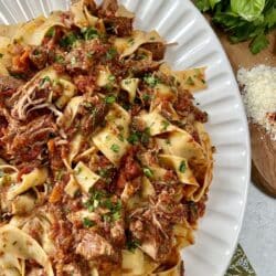 Finished pappardelle with pork ragu recipe in pan on white platter with fresh herbs, grated cheese and wooden spoon in background.