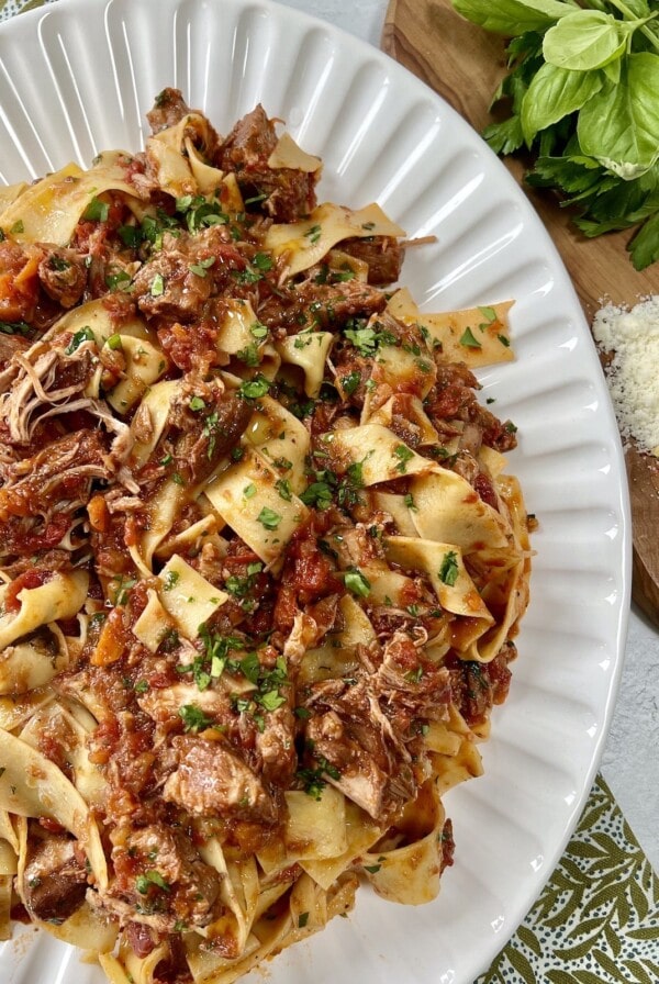 Finished pappardelle with pork ragu recipe in pan on white platter with fresh herbs, grated cheese and wooden spoon in background.