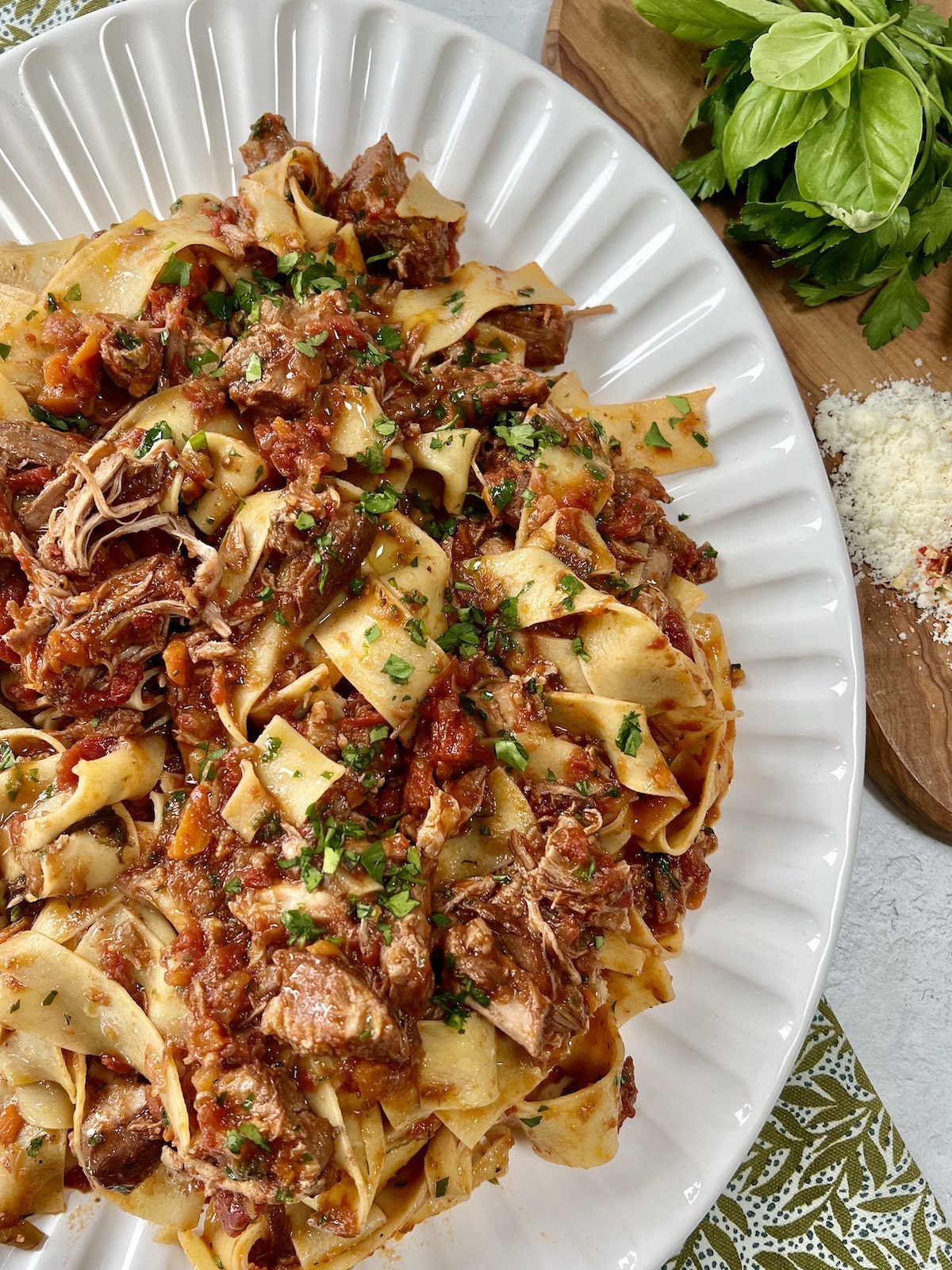 Finished pappardelle with pork ragu recipe in pan on white platter with fresh herbs, grated cheese and wooden spoon in background.