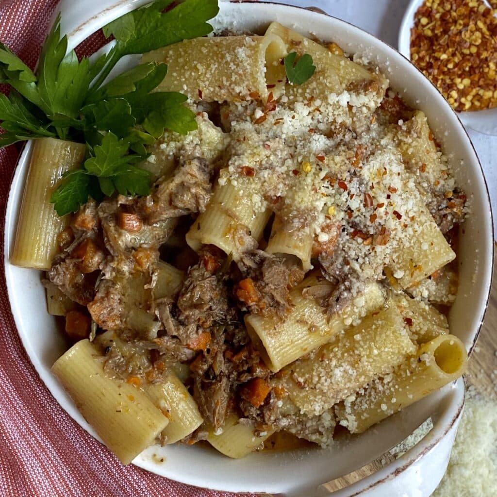 Wild boar bolognese with rigatoni pasta in white dish with parsley garnish on red towel with crushed red pepper in background.