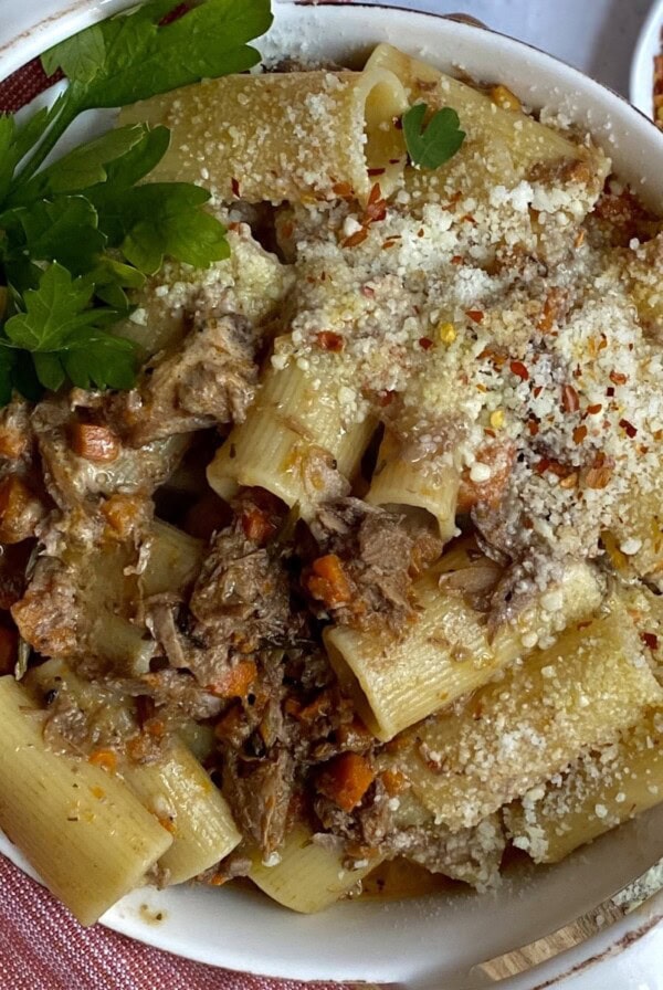 Wild boar bolognese with rigatoni pasta in white dish with parsley garnish on red towel with crushed red pepper in background.