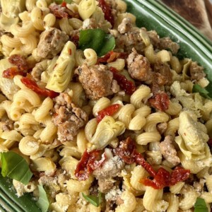 Artichoke sausage pasta in green bowl with wooden spoon in background.