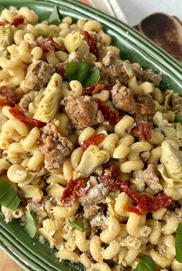Artichoke sausage pasta in green bowl with wooden spoon in background.