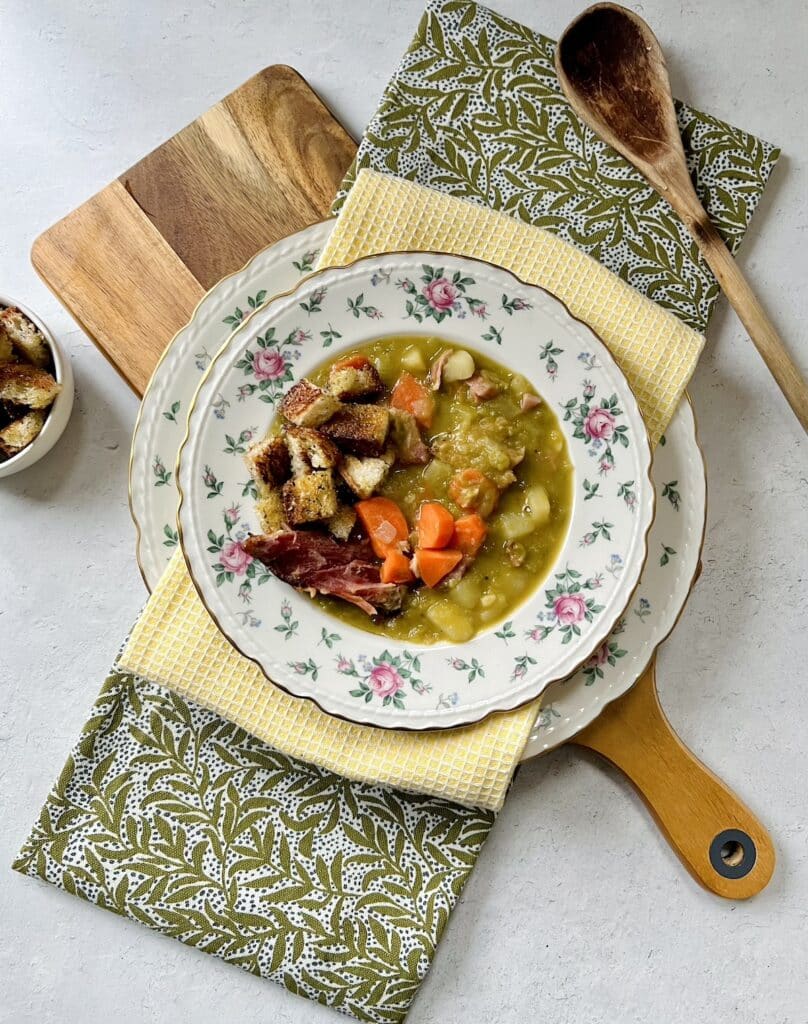 Split Pea and Ham Soup with Potatoes in decorative bowl styled on yellow and green towels on a cutting board with wooden spoon and dish of croutons surrounding it.