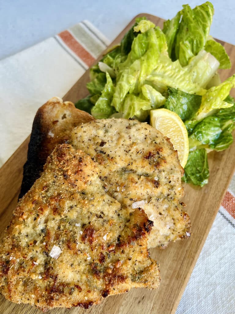 Crispy baked chicken cutlets sprinkled with sea salt and serving board with green salad and lemon wedge.