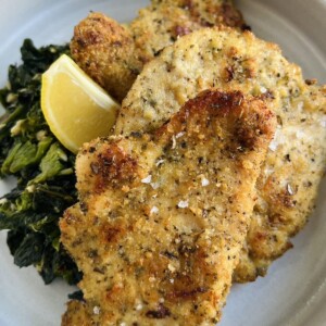 Baked chicken cutlets, Italian style, on plate with saut&eacute;ed broccoli rabe and lemon wedge.