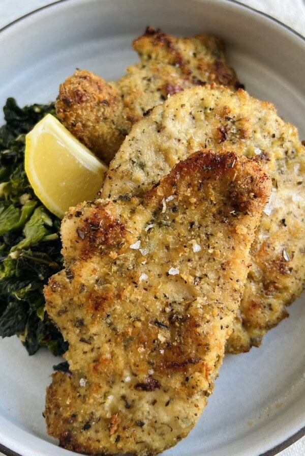 Baked chicken cutlets, Italian style, on plate with sautéed broccoli rabe and lemon wedge.