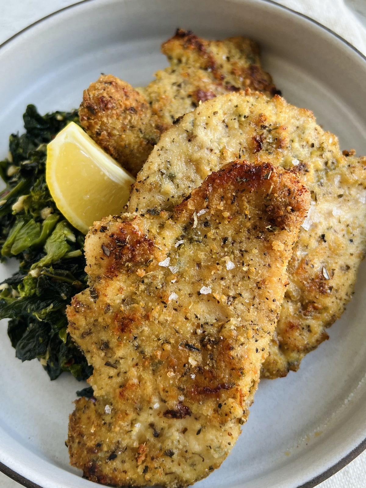 Baked chicken cutlets, Italian style, on plate with sautéed broccoli rabe and lemon wedge.