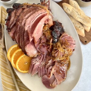 Overhead view of Dutch oven baked ham with marsala glaze plated on an oval platter with orange slices and onions, with mustard, cranberry sauce, additional glaze and bread in the background.