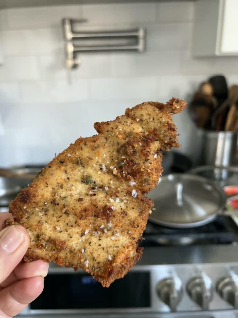Close up of one breaded chicken cutlet, Italian style, being held with the stove in the background.