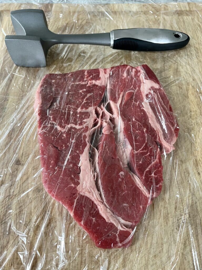 Piece of chuck steak in between plastic wrap with meat mallet.