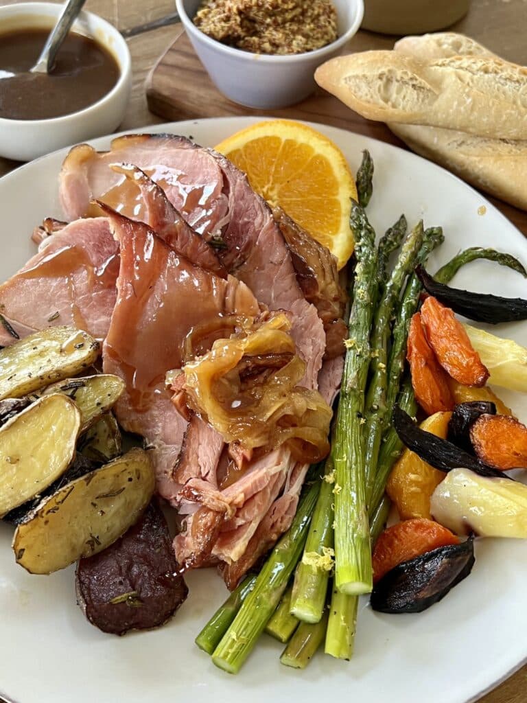 Baked spiral ham plated on a white dinner plate with asparagus, potatoes, carrots and orange slices, with mustard, additional glaze and bread in background.