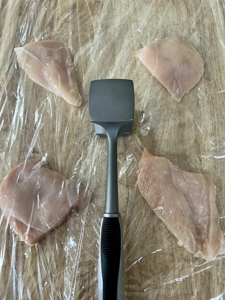 Four pieces of thinly sliced chicken breast in between plastic wrap ready to be pounded out with a meat tenderizer, which is also pictured.