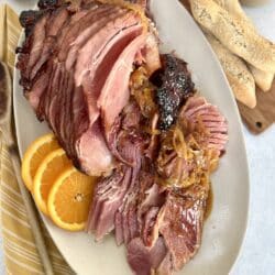 Overhead view of Dutch oven baked ham with marsala glaze plated on an oval platter with orange slices and onions, with mustard, cranberry sauce, additional glaze and bread in the background.