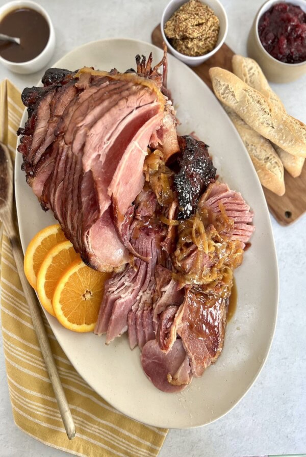 Overhead view of Dutch oven baked ham with marsala glaze plated on an oval platter with orange slices and onions, with mustard, cranberry sauce, additional glaze and bread in the background.