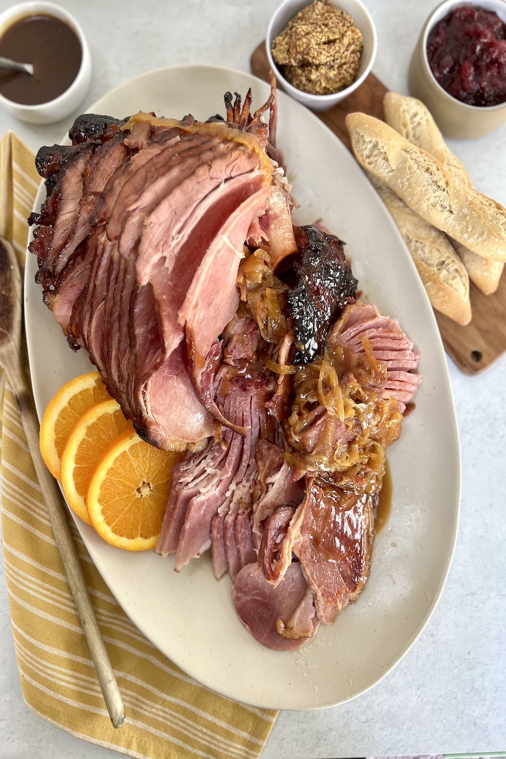 Overhead view of Dutch oven baked ham with marsala glaze plated on an oval platter with orange slices and onions, with mustard, cranberry sauce, additional glaze and bread in the background.