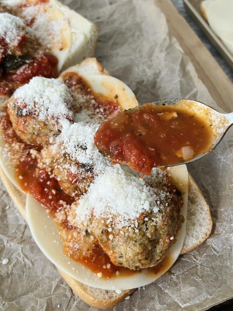 Building a meatball parm sub with turkey pesto meatballs, tomato basil sauce, provolone and pecorino cheese on a long roll.