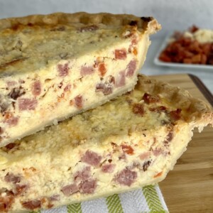 Two wedges of Italian Easter Meat Pie on decorative towel on cutting board with small dish of prepped meats and cheeses in background.