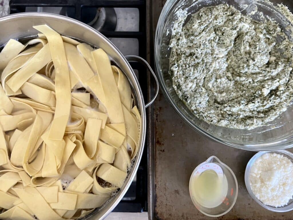 Pasta cooking in pot of boiling water next to bowls of pesto ricotta mixture, lemon juice and grated cheese.