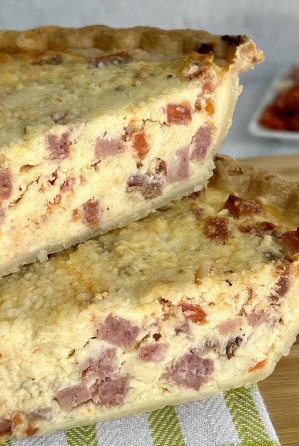 Two wedges of Italian Easter Meat Pie on decorative towel on cutting board with small dish of prepped meats and cheeses in background.