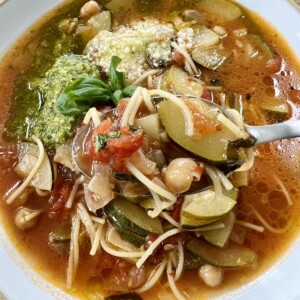 Italian zucchini soup in white bowl with basil pesto, grated cheese and basil sprig garnish with spoon in bowl.