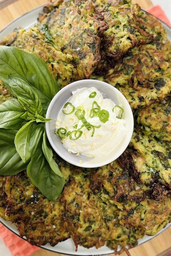 Italian zucchini fritters on round white plated shingled around a small bowl of sour cream topped with lemon zest and sliced green onion, with a basil sprig garnish.