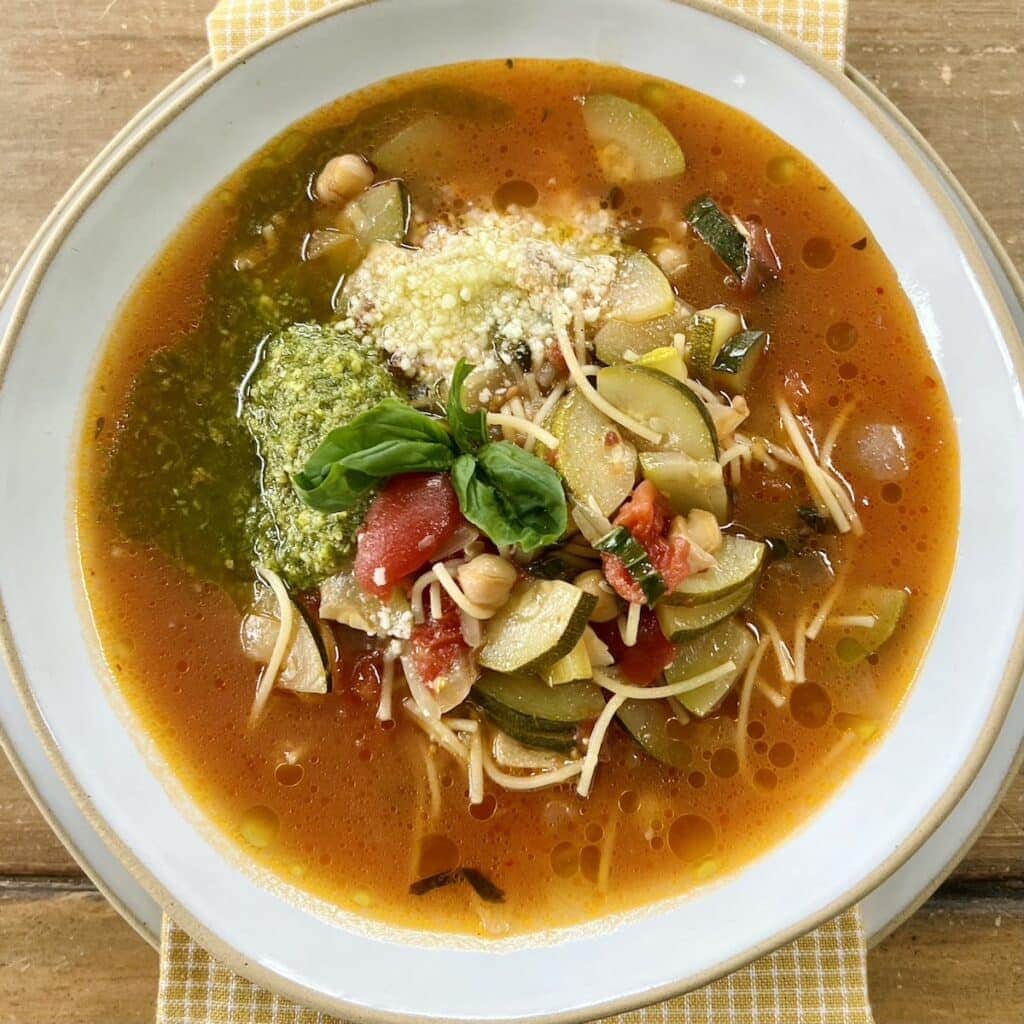 Italian zucchini soup in white bowl with basil pesto, grated cheese and basil sprig garnish, over a yellow towel.