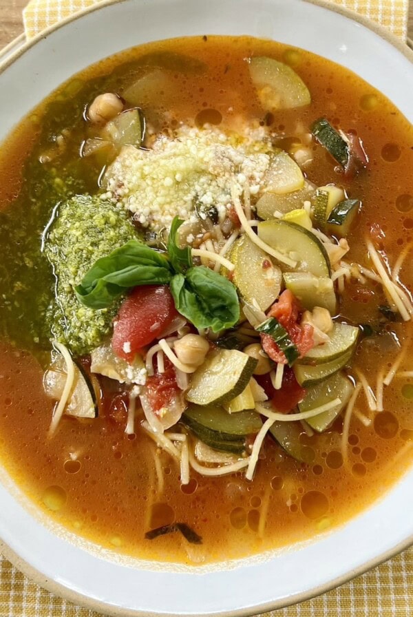 Italian zucchini soup in white bowl with basil pesto, grated cheese and basil sprig garnish, over a yellow towel.