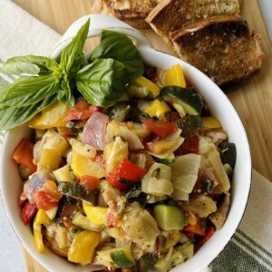 Giambotta Italian vegetable stew in white dish on towel on cutting board with basil garnish and toasted bread next to it.