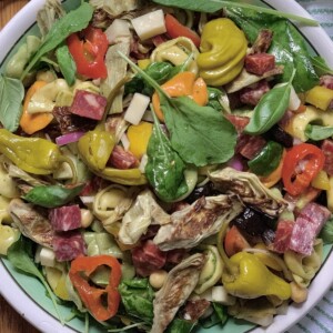 Antipasto tortellini pasta salad in decorative bowl on a white and green towel with pickled peppers in background.