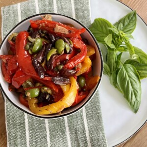 Beautiful bowl of Italian peperonata in white bowl with black rim over green towel over white plate with basil garnish.